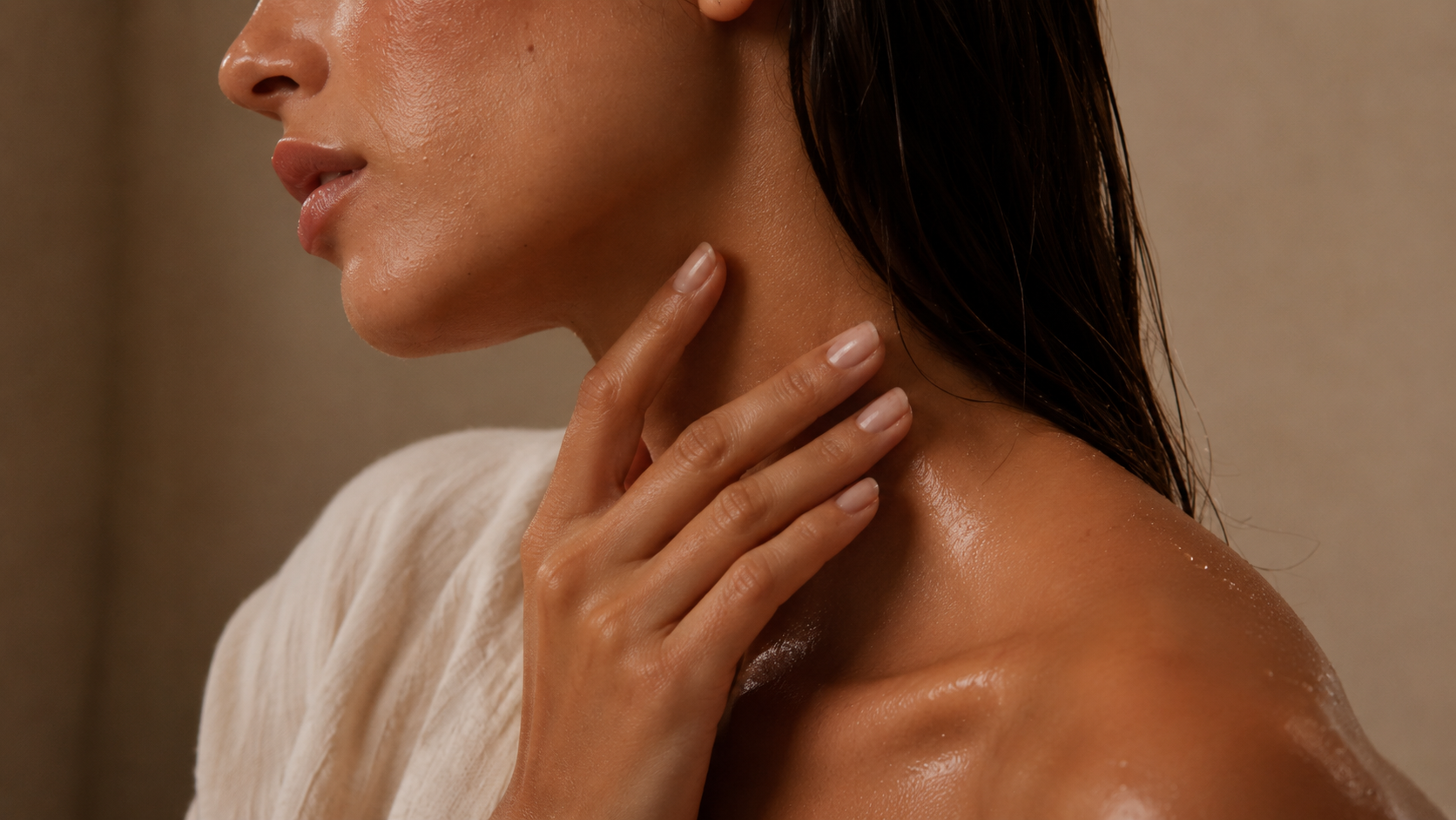 A hand smooths body oil along the neck and collarbone on slightly damp skin after a shower.