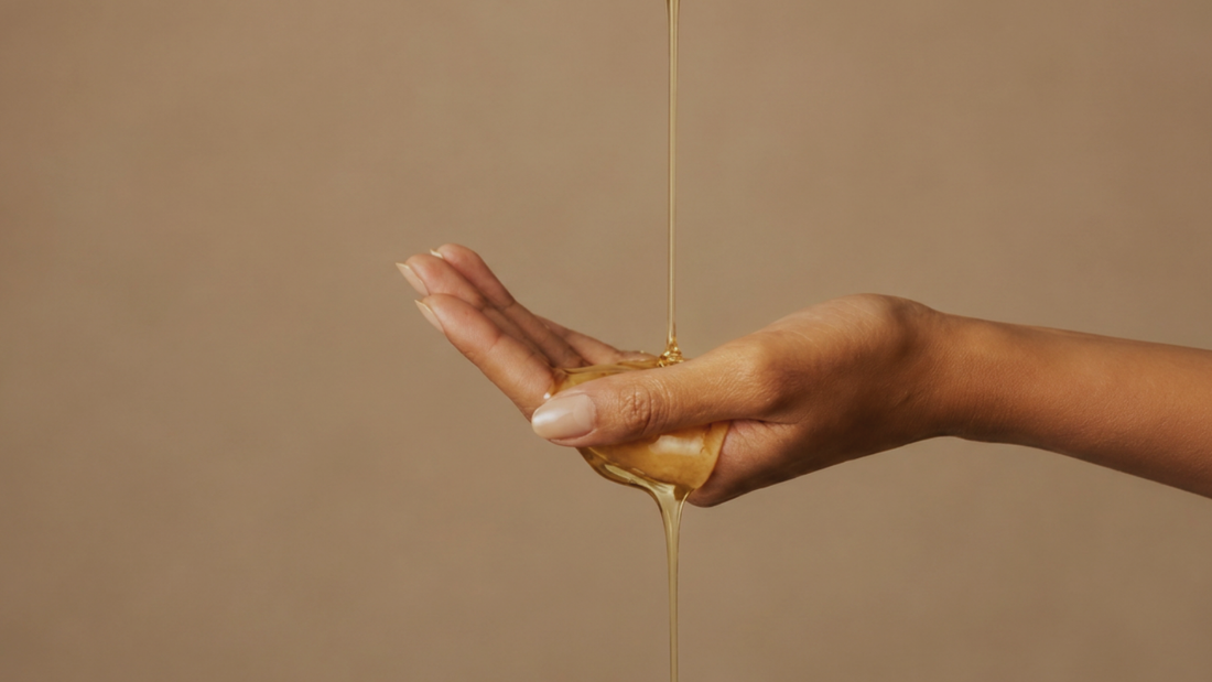 Golden body oil drips over an open palm against a soft, light background, showing a dry-oil texture.