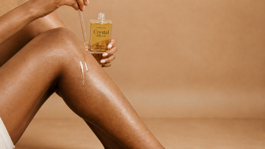 A hand holds Crystal Body Oil beside a glowing, freshly oiled leg, a soft dewy finish.