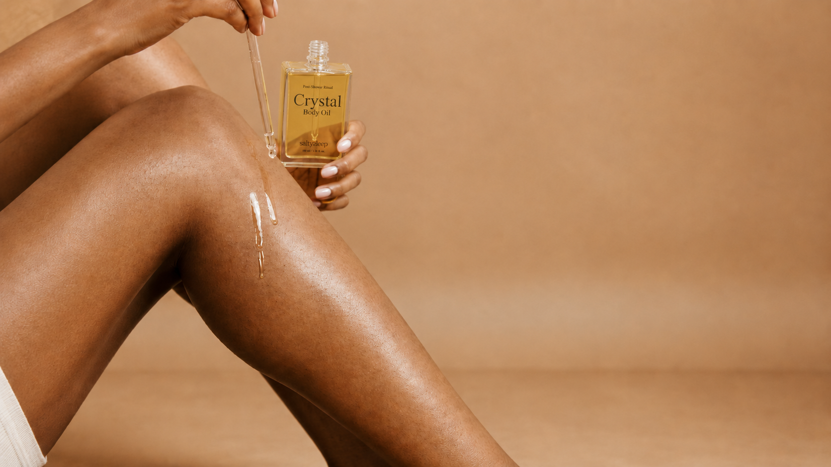 A hand holds Crystal Body Oil beside a glowing, freshly oiled leg, a soft dewy finish.