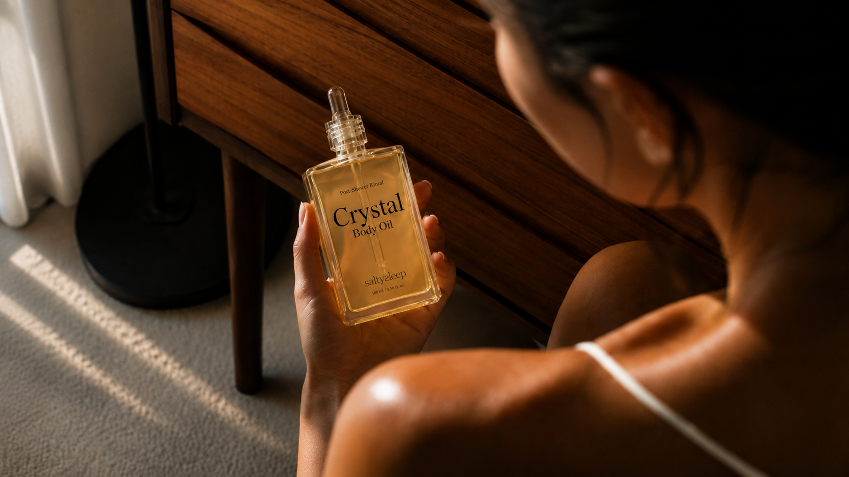 Crystal Body Oil resting on soft silk fabric in warm light before bed.