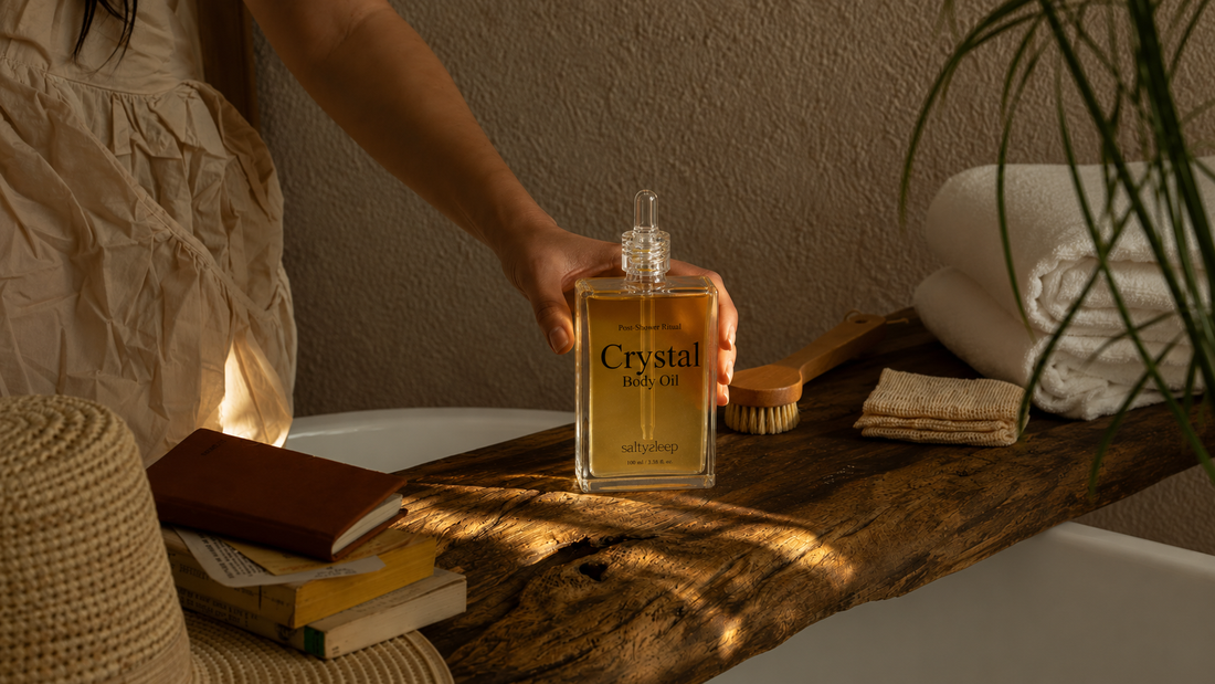 Crystal Body Oil on a bath tray beside a book, a calm warm-lit post-shower still life.